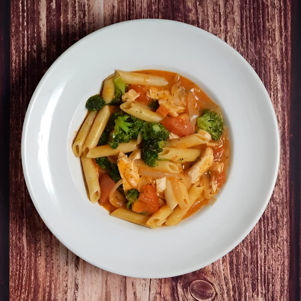 Penne pasta with chicken, broccoli, and tomatoes in a creamy orange sauce on a white plate.