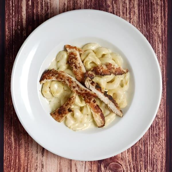 Creamy pasta with grilled chicken strips in a white bowl on a wooden table.
